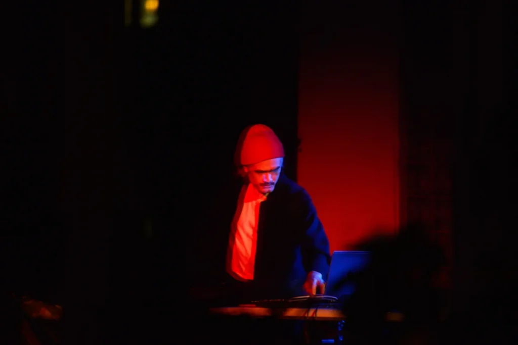 picture shows Eric Wong standing over a turntable in a dark room. He is wearing a white shirt, a dark jacket and a red beanie. He is partially lit by red light